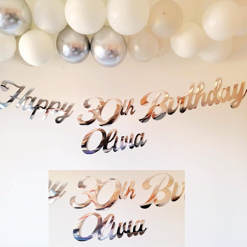 May include: Silver mirror-like Happy 30th Birthday Olivia banner. The banner is made of individual letters and numbers. The banner is hanging on a white wall with a silver and white balloon garland above it.
