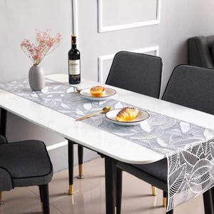 May include: A white table runner with a grey leaf pattern on a white marble table with two chairs and a bottle of wine.