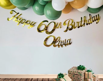 Op de afbeelding: Gouden folie Happy 60th Birthday banner met de naam Olivia, groene en gouden ballonnen en bruine cadeaudozen met groen lint.