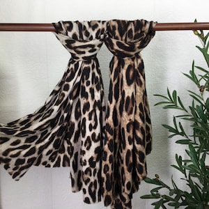 May include: Two leopard print scarves are draped over a wooden rail. The scarves have a pattern of black spots on a tan and cream background. The fabric is lightweight and flowing, suggesting a soft texture. The scarves are displayed against a white background.