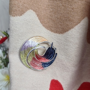 May include: A silver brooch with a multi-coloured design. The brooch is made of two overlapping circles, one with a green, yellow, and purple design and the other with a red, black, and blue design.