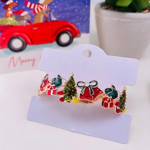 May include: A gold-coloured Christmas bracelet with red bells, green Christmas trees, and wrapped gifts. The bracelet is attached to a white card, with a festive card in the background.