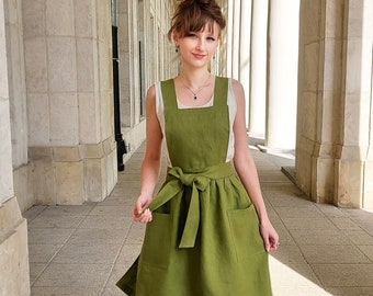Puede incluir: Una mujer lleva un vestido de delantal de lino verde con una blusa blanca debajo. El vestido tiene un cinturón ancho y dos bolsillos grandes. Está de pie en un pasillo con columnas blancas.