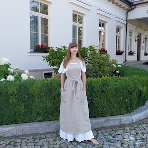 Może przedstawiać: Kobieta w długim, jasnobrązowym lnianym fartuchu z kieszeniami i paskiem do wiązania. Stoi przed białym budynkiem z oknami i zielonymi krzewami.