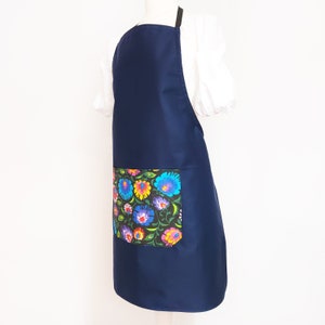 K&ouml;nnte beinhalten: Eine marineblaue Sch&uuml;rze mit einer gro&szlig;en Tasche mit Blumendruck. Die Tasche hat einen schwarzen Hintergrund mit bunten Blumen und gr&uuml;nen Bl&auml;ttern.