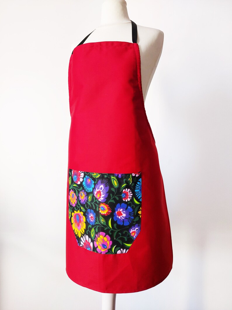 K&ouml;nnte beinhalten: Rote Sch&uuml;rze mit einer gro&szlig;en Tasche mit Blumendruck. Die Sch&uuml;rze hat schwarze Tr&auml;ger.