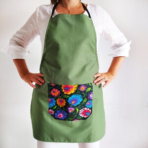 K&ouml;nnte beinhalten: Eine gr&uuml;ne Sch&uuml;rze mit einer gro&szlig;en Tasche, die mit einem bunten Blumenmuster verziert ist. Die Sch&uuml;rze hat verstellbare Tr&auml;ger und eine lange Sch&uuml;rze.