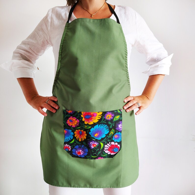 K&ouml;nnte beinhalten: Eine gr&uuml;ne Sch&uuml;rze mit einer gro&szlig;en Tasche, die mit einem bunten Blumenmuster verziert ist. Die Sch&uuml;rze hat verstellbare Tr&auml;ger und eine lange Sch&uuml;rze.