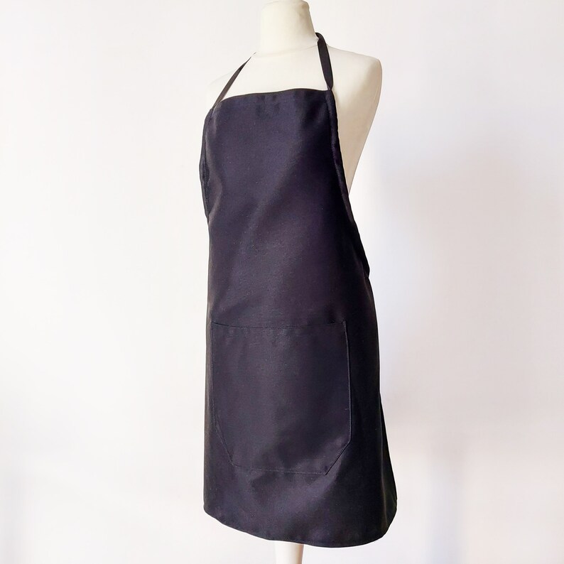 K&ouml;nnte beinhalten: Eine schwarze Sch&uuml;rze mit einer gro&szlig;en Tasche auf der Vorderseite. Die Sch&uuml;rze ist aus glattem, strapazierf&auml;higem Stoff.