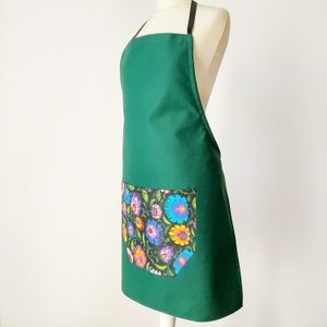 K&ouml;nnte beinhalten: Eine dunkelgr&uuml;ne Sch&uuml;rze mit einer gro&szlig;en Tasche mit Blumendruck. Die Tasche hat einen schwarzen Hintergrund mit bunten Blumen und Bl&auml;ttern.