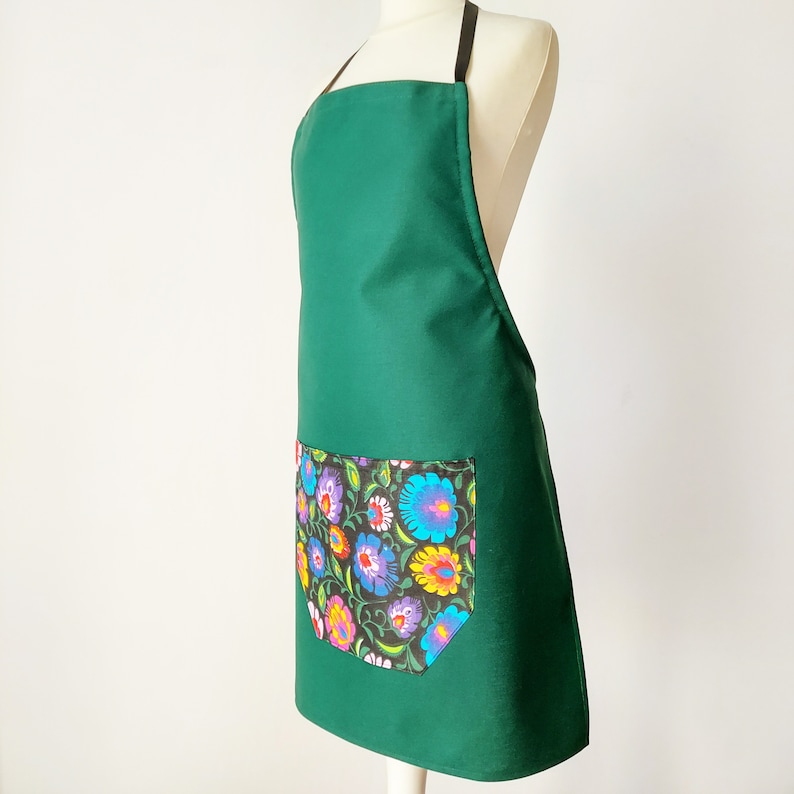 K&ouml;nnte beinhalten: Eine dunkelgr&uuml;ne Sch&uuml;rze mit einer gro&szlig;en Tasche mit Blumendruck. Die Tasche hat einen schwarzen Hintergrund mit bunten Blumen und Bl&auml;ttern.