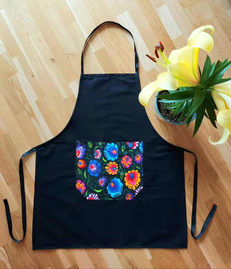 K&ouml;nnte beinhalten: Schwarze Sch&uuml;rze mit einer gro&szlig;en Tasche mit einem bunten Blumenmuster. Die Sch&uuml;rze hat verstellbare Tr&auml;ger.