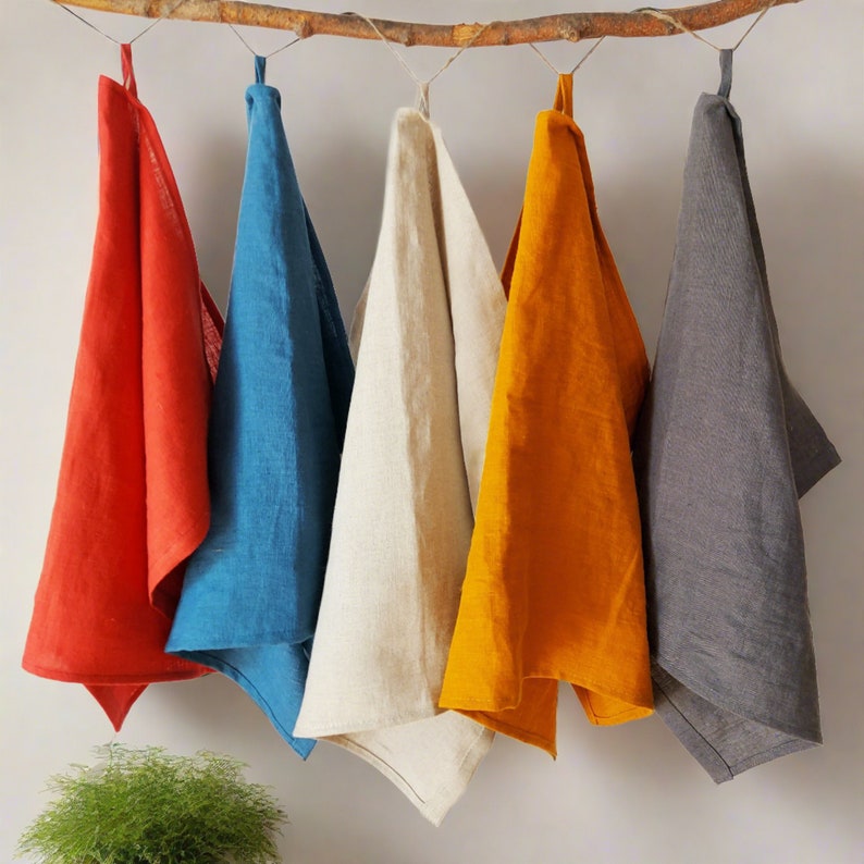 May include: Five linen tea towels in different colors: red, blue, cream, orange, and gray. The towels are hanging from a wooden branch.