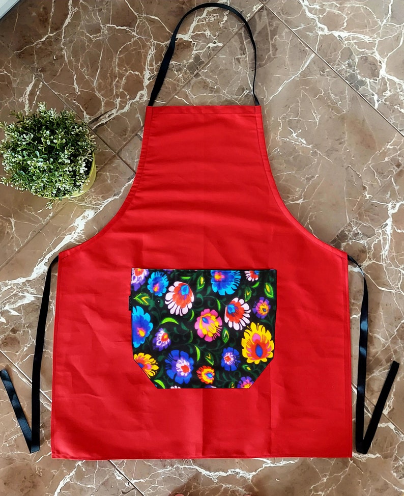 K&ouml;nnte beinhalten: Rote K&uuml;chensch&uuml;rze mit einer schwarzen Tasche mit Blumendruck. Die Sch&uuml;rze hat verstellbare schwarze Tr&auml;ger.