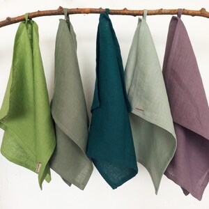 May include: Five linen tea towels hanging on a branch. The towels are in shades of green, teal, and purple.