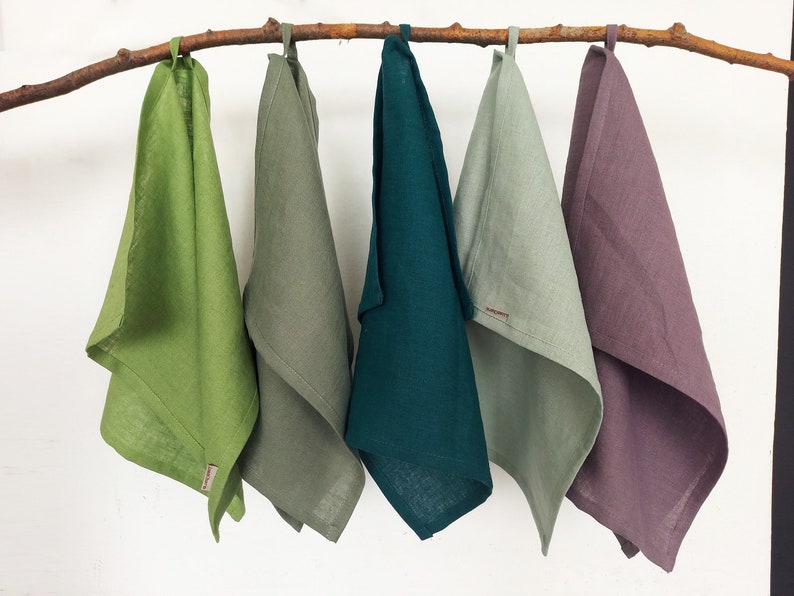May include: Five linen tea towels hanging on a branch. The towels are in shades of green, teal, and purple.