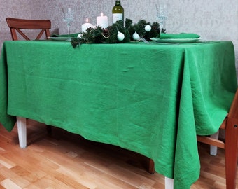 May include: A green linen tablecloth with a white wooden table and a green garland with white ornaments. The table is set with two glasses, a bottle of wine, and two candles.