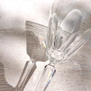 May include: A clear crystal wine glass with a long stem and a detailed, textured bowl. The glass is lying on its side, casting a shadow. The image is lit with natural light, creating a rainbow effect.