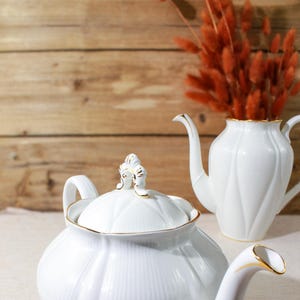 Puede incluir: Tetera de porcelana blanca con ribete dorado y diseño floral. La tetera tiene una tapa con un pomo decorativo. La tetera está sobre un mantel de lino blanco.