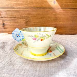 May include: A vintage teacup and saucer set with a floral design. The cup has a blue flower handle and the saucer has a yellow rim.