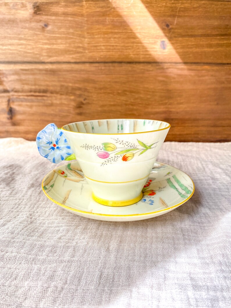 May include: A vintage teacup and saucer set with a floral design. The cup has a blue flower handle and the saucer has a yellow rim.