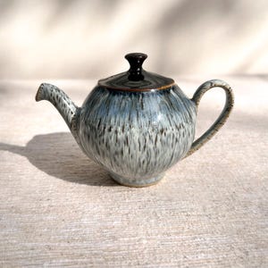 以下が含まれることがあります： 青とグレーの斑点模様の釉薬と黒い蓋が付いたセラミックティーポット。 ティーポットには、湾曲したハンドルと注ぎ口があります。 ティーポットはニュートラルカラーの表面に置かれています。