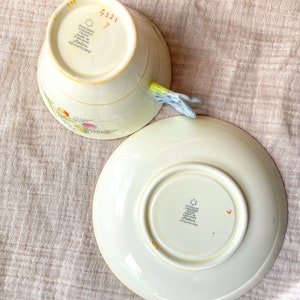 May include: A vintage teacup and saucer set with a floral design. The teacup has a handle shaped like a flower. The bottom of the teacup and saucer both have the words "Paragon Fine China Made in England" and a star.