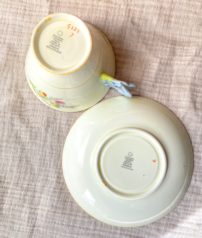 May include: A vintage teacup and saucer set with a floral design. The teacup has a handle shaped like a flower. The bottom of the teacup and saucer both have the words "Paragon Fine China Made in England" and a star.