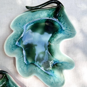 May include: A ceramic dish with an irregular, organic shape featuring a glossy glaze in shades of deep forest green, teal, and pale mint. The center has a dark, saturated pool of color that transitions into lighter, mottled edges.