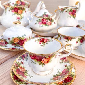 May include: A vintage tea set featuring a teapot, sugar bowl, creamer, and teacups with saucers. The white porcelain set is adorned with a floral rose pattern in shades of red, pink, and yellow, with gold trim.