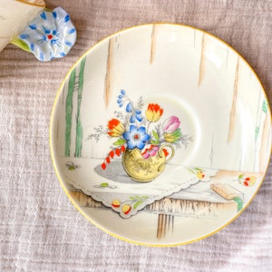 May include: A white ceramic saucer with a gold rim features a painted design of a bouquet of flowers in a vase. The flowers are red, orange, yellow, blue, and green. The saucer has a scalloped edge and a delicate floral pattern.