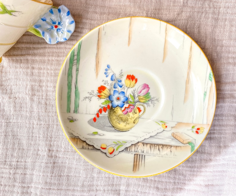 May include: A white ceramic saucer with a gold rim features a painted design of a bouquet of flowers in a vase. The flowers are red, orange, yellow, blue, and green. The saucer has a scalloped edge and a delicate floral pattern.