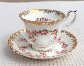 Puede incluir: Juego de taza y platillo blancos con un diseño floral de rosas rosas y ribete dorado. La taza tiene un asa curvada y descansa sobre un platillo a juego. El conjunto está decorado con rosas rosas y hojas verdes.