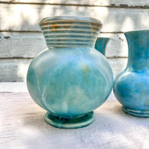 May include: Three ceramic vases in shades of turquoise and blue. The central vase has a bulbous body, a ribbed neck, and a small base. The other two vases have similar shapes, with variations in the neck and body design. The vases are on a white surface.