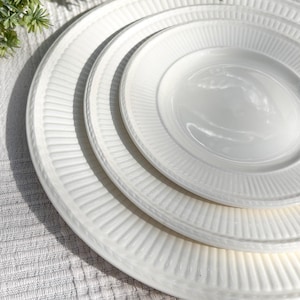 May include: A stack of three white ceramic plates with a ribbed design. The plates are of varying sizes, with the largest at the bottom and the smallest at the top. The plates are arranged on a white textured surface.
