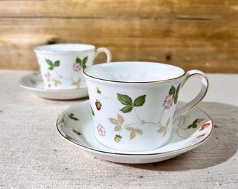 Puede incluir: Dos tazas de té blancas y platillos con un patrón floral que presenta fresas y hojas. Las tazas tienen un borde dorado y los platillos tienen una banda dorada alrededor del borde.