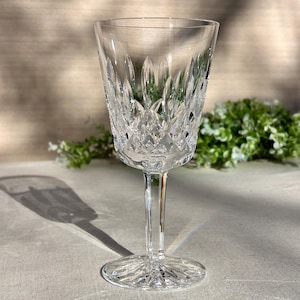May include: A clear crystal wine glass with a detailed cut pattern. The glass has a long stem and a round base. The cut pattern includes vertical lines and diamond shapes. The glass is empty and sits on a light grey surface.