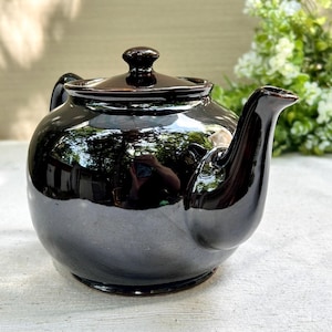 May include: A glossy, dark brown ceramic teapot with a rounded body, a curved spout, and a matching lid with a small knob handle. The surface of the teapot is highly reflective, showing a clear mirror-like finish that captures light and surroundings.