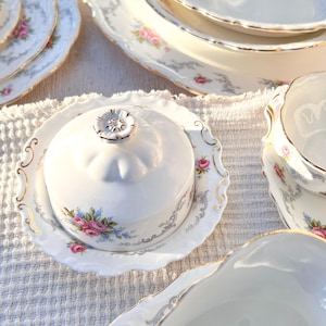 Puede incluir: Un conjunto de vajilla de porcelana blanca con un diseño floral de rosas y ribetes dorados. El juego incluye platos, cuencos y una mantequera con tapa. Los platos están colocados sobre una superficie blanca texturizada.