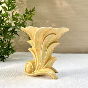 Peut inclure: Un vase en céramique présentant une silhouette évasée en forme d'éventail avec des bords incurvés et organiques. La surface présente un émail marbré dans des tons jaune pâle, crème et vert doux, avec une finition brillante soulignant les reliefs.