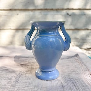 Peut inclure: Un vase en céramique bleu clair avec deux anses incurvées et une base en piédestal. Le vase a une finition lisse et brillante et un bord légèrement évasé. Le fond est une surface en bois blanc vieilli.
