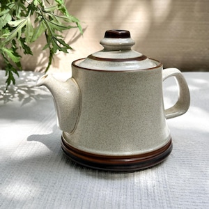 以下が含まれることがあります： 斑点のあるオフホワイト仕上げの陶器製ティーポット。蓋にはダークブラウンの縁取りがあり、同色のダークブラウンの底面と湾曲したハンドルが特徴です。ティーポットには短く先細りの注ぎ口があり、幅広で平らな底面を備えています。