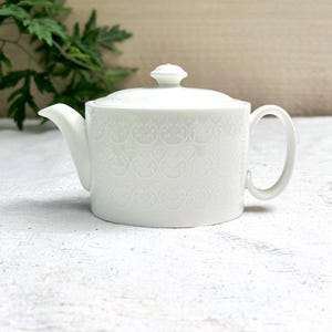 Peut inclure: Une théière en céramique blanche dotée d'un corps cylindrique avec un motif floral répétitif en relief, un couvercle assorti avec un petit bouton, un bec verseur incurvé et une anse en forme de C sur le côté, au fini propre et lumineux.