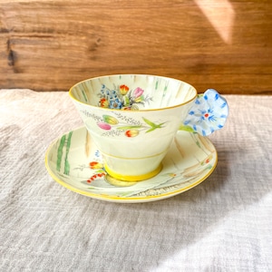 May include: A vintage teacup and saucer set with a floral design. The cup has a yellow rim and a blue flower handle. The saucer has a yellow rim and a floral design.