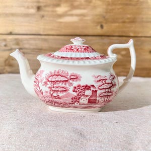 May include: A white teapot with a pink floral design and a lid. The teapot has a spout and a handle. The teapot is sitting on a white tablecloth.