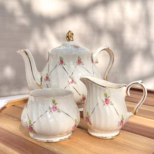 Puede incluir: Un juego de té de porcelana blanca con tetera, jarra de crema y azucarero. Cada pieza está decorada con rosas rosas, ribetes dorados y un patrón de diamantes. La tetera tiene un adorno y asa dorados.