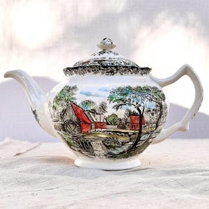May include: A white ceramic teapot with a lid, decorated with a rural landscape scene including red buildings and trees. The handle and spout are white, and the lid has a black and white floral design.
