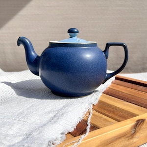 Puede incluir: Una tetera de cerámica azul con tapa y asa a juego. La tetera tiene un cuerpo redondeado y un pico en el lado izquierdo. La tapa es azul claro en la parte superior y azul oscuro en los lados. La tetera está sobre un paño blanco y una bandeja de madera.