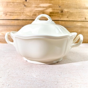 Puede incluir: Una sopera de cerámica blanca con tapa y asas. La sopera tiene un borde festoneado y un asa de bucle en la tapa. El fondo es de madera clara.