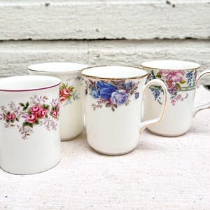 May include: Four white ceramic mugs with floral patterns. One mug displays pink roses, another blue roses, and the others assorted flowers. Each mug has a gold rim and handle, set against a white backdrop.